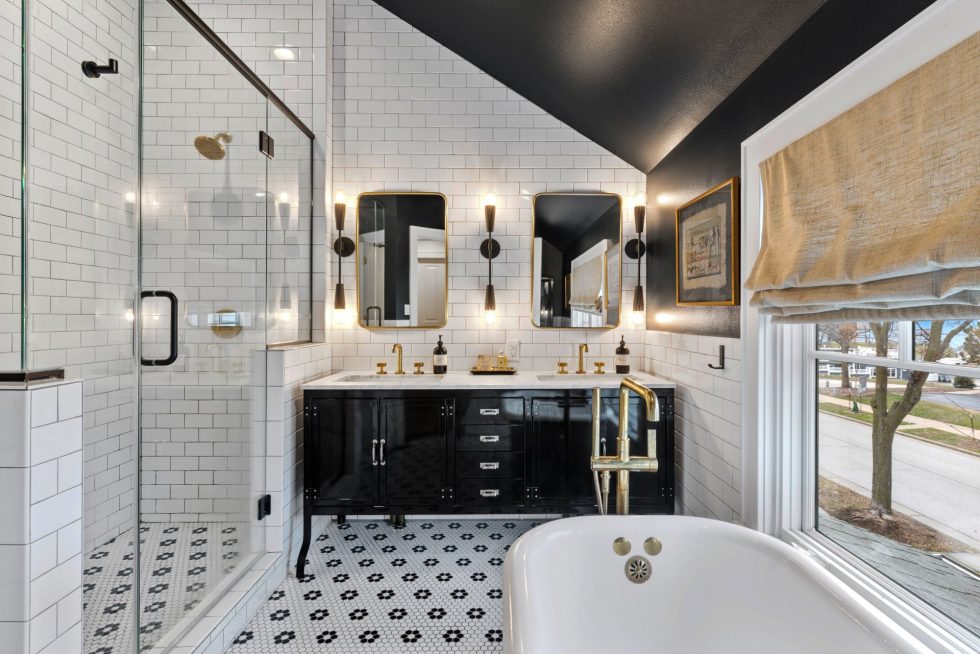 A modern bathroom by Milwaukee Home Remodeling features a walk-in glass shower, a double sink vanity with black cabinets, and gold fixtures. White subway tiles and patterned black and white floor tiles are present throughout. A large window has a rolled-up beige shade, and a freestanding tub is in the foreground.
