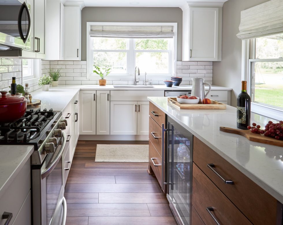 A modern kitchen with white cabinets, stainless steel appliances, and hardwood floors boasts a sleek design. The countertops are white, and there's a large window above the sink. A wooden tray with fruits and a bottle of wine sits on the island featuring a wine cooler—a true masterpiece by an award-winning remodeling firm.