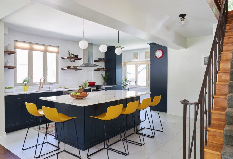 A modern kitchen designed by Milwaukee Remodeling Contractor: Design Tech Remodeling, featuring a large island with a white marble countertop and yellow bar stools. Navy blue cabinets and open wooden shelves line the walls. Three pendant lights hang above the island, and a staircase with wooden steps is visible on the right.