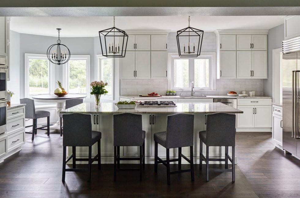 A luxury kitchen featuring a large island with a marble countertop and four dark grey bar stools. Two geometric pendant lights hang above the island. The kitchen includes white cabinets, stainless steel appliances, and a dining nook with a round table and chairs—perfect for any high-end remodeling project.