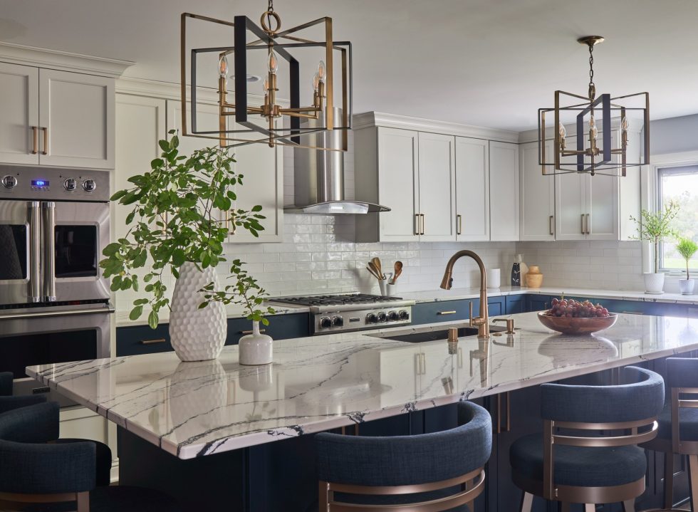 Modern kitchen with white cabinets and navy blue island. Marble countertop features a sink and vase with greenery. Two geometric pendant lights hang above. Stainless steel appliances, including double ovens and a range hood, are built into the cabinetry.