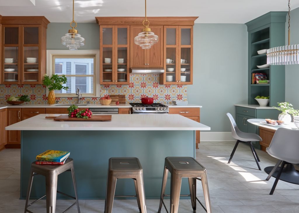 A modern kitchen with wooden cabinets, colorful patterned backsplash, and teal-colored island. The island has three metal stools and features a white countertop with strawberries and a cutting board. The space is lit by pendant lights and has a small dining area.